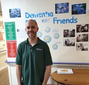 Man wearing a Rushcliffe Care uniform posed in front of a wall display that says 'Dementia Friends'